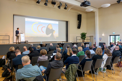 Referent auf Bühne, davor drei Stuhlreihen besetzt mit Zuhörenden