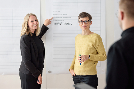 Arbeitssituation mit drei Personen: Eine Mitarbeiterin zeigt auf ein Plakat an der Wand.
