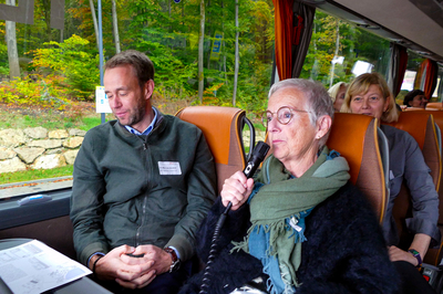 2 Personen sitzen im Bus; die Frau spricht ins Mikrofon
