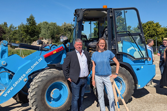 Zwei Personen stehen vor einem Radlader, eine von ihnen hält einen Spaten in der Hand